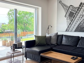 A modern living room with a black leather sofa, a wooden coffee table, a floor lamp, and a large window with a view of a tree and fence.