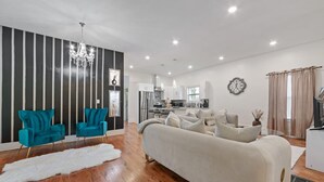 Ein modernes Wohnzimmer mit einer Couch, zwei Sesseln, einer Chandelieber und einer Küche im Hintergrund.