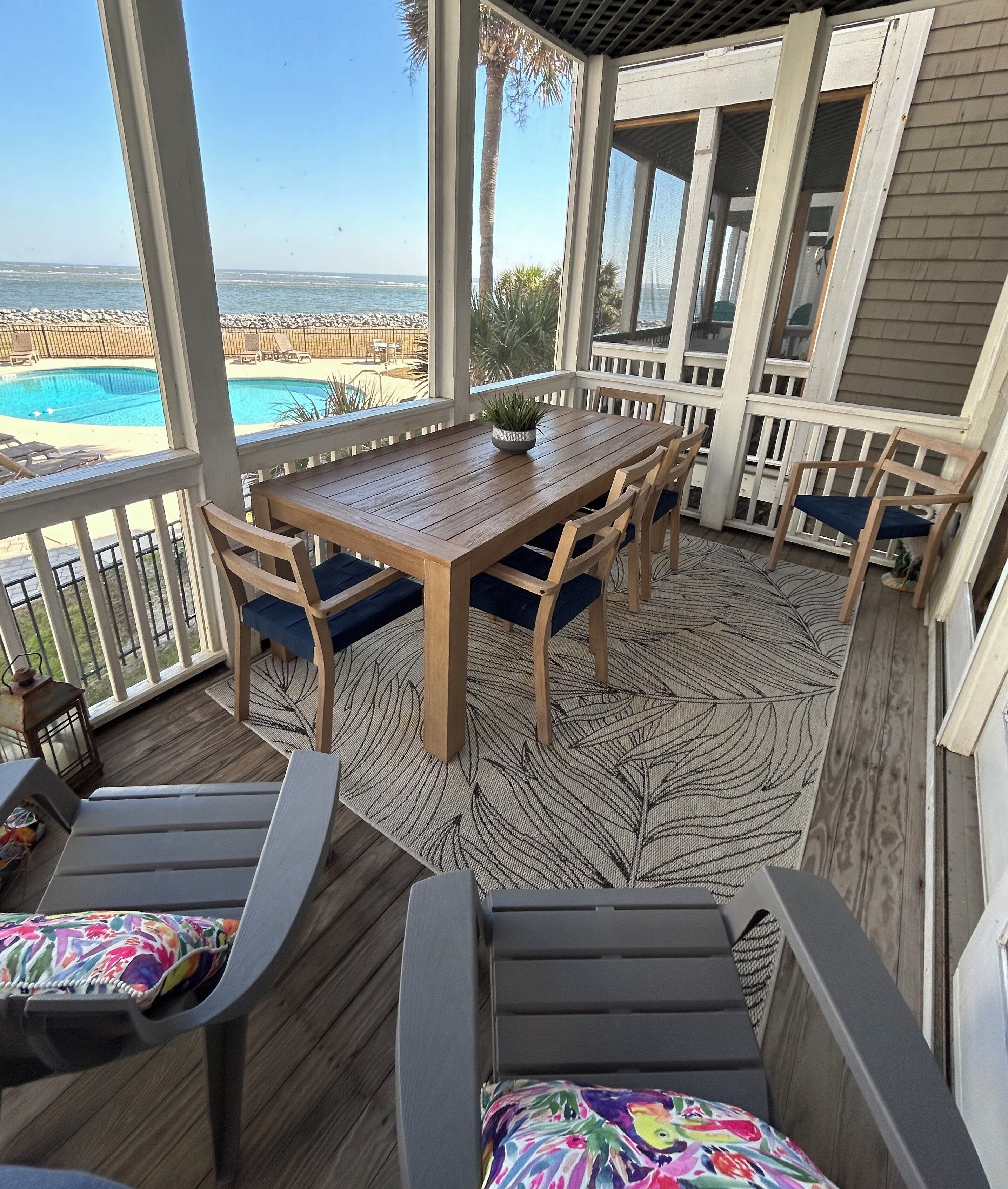 Un patio techado al aire libre con una mesa y sillas de madera, una piscina al fondo y vista al océano.