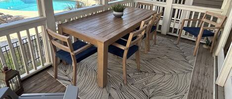 Un patio techado al aire libre con una mesa y sillas de madera, una piscina al fondo y vista al océano.