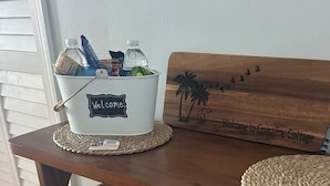 A wooden table with a woven placemat, a white bucket labeled "Welcome," and a wooden sign with palm trees and the text "Welcome to Coastal Cottage."