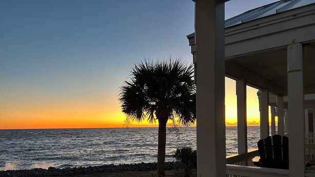 A poolside view with a sunset over the ocean, a palm tree, and a deck with chairs.