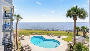 A pool area with a view of the ocean, lounge chairs, and a building with balconies.