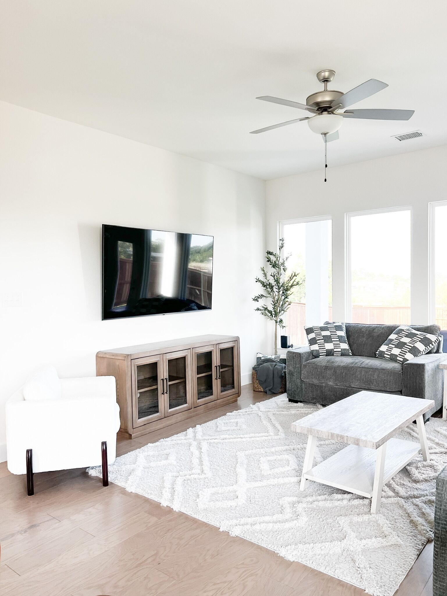 A modern living room with a flat-screen TV, a ceiling fan, a wooden cabinet, a grey sofa, and a white coffee table.