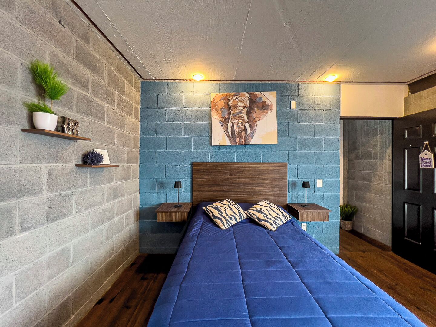 Un dormitorio con pared de ladrillo azul, cabecera de madera, cama con ropa de cama azul, dos mesitas de noche con lámparas y una puerta con un cartel.