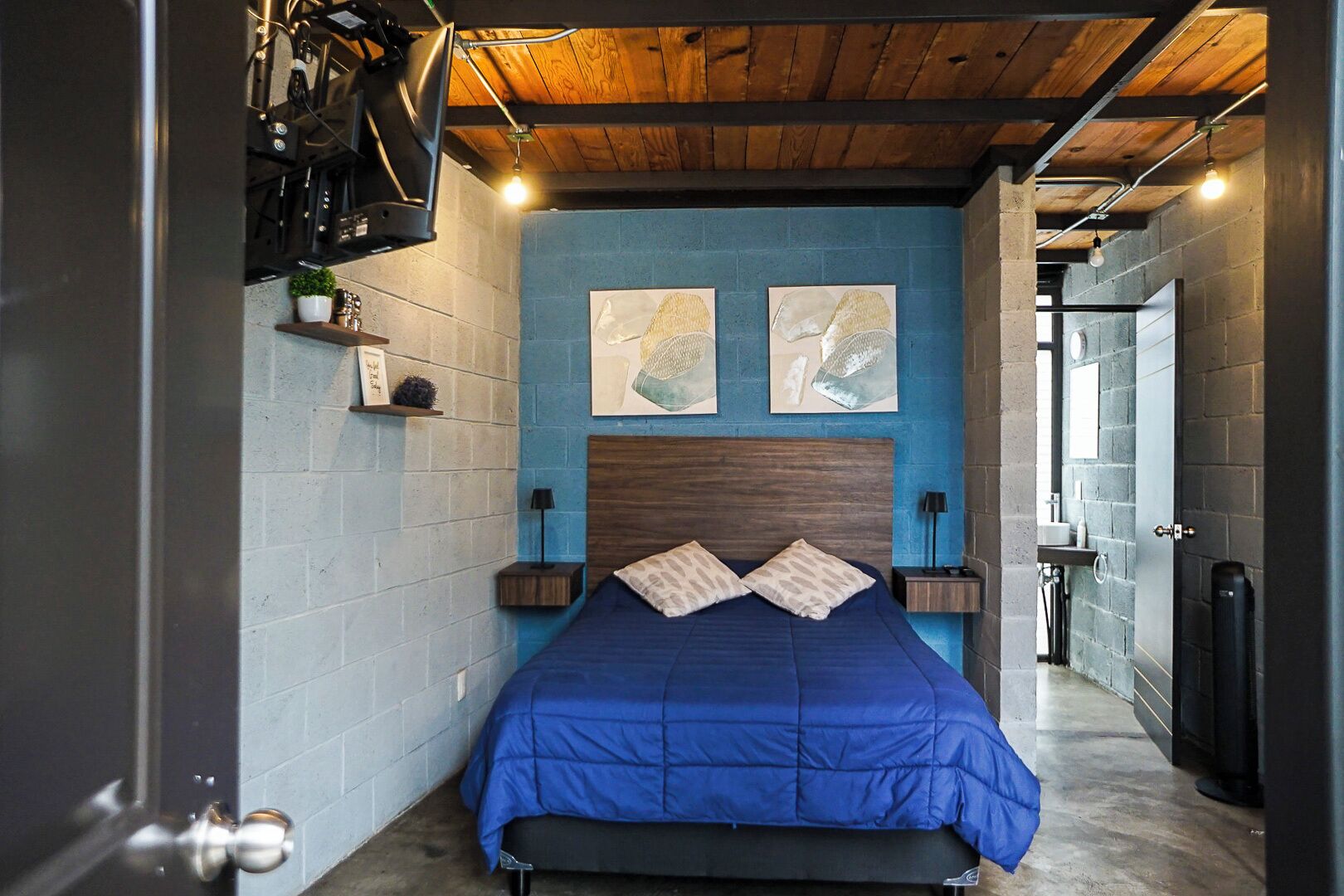 Quarto com parede azul, teto de madeira e cama com cobre azul. Há duas pinturas na parede e uma pequena estante com uma planta.