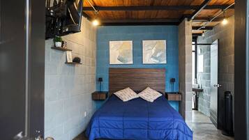 Quarto com parede azul, teto de madeira e cama com cobre azul. Há duas pinturas na parede e uma pequena estante com uma planta.