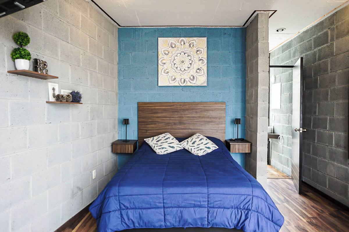 Un dormitorio con pared azul, cabecera de madera y cama grande con ropa de cama azul. Hay dos mesitas de noche con lámparas, un adorno en la pared y una puerta a la derecha.