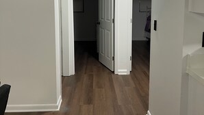 Un pasillo con suelo de madera, paredes blancas y una puerta que da a otra habitación.