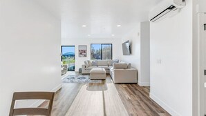 A modern living room with a large sectional sofa, a flat-screen TV mounted on the wall, and large windows offering a view of the outdoors. - High-end, convenient, new build! (Juneau)