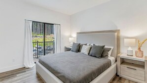 A bedroom with a large bed, a nightstand with a lamp, and a view of a balcony with outdoor furniture. - High-end, convenient, new build! (Juneau)