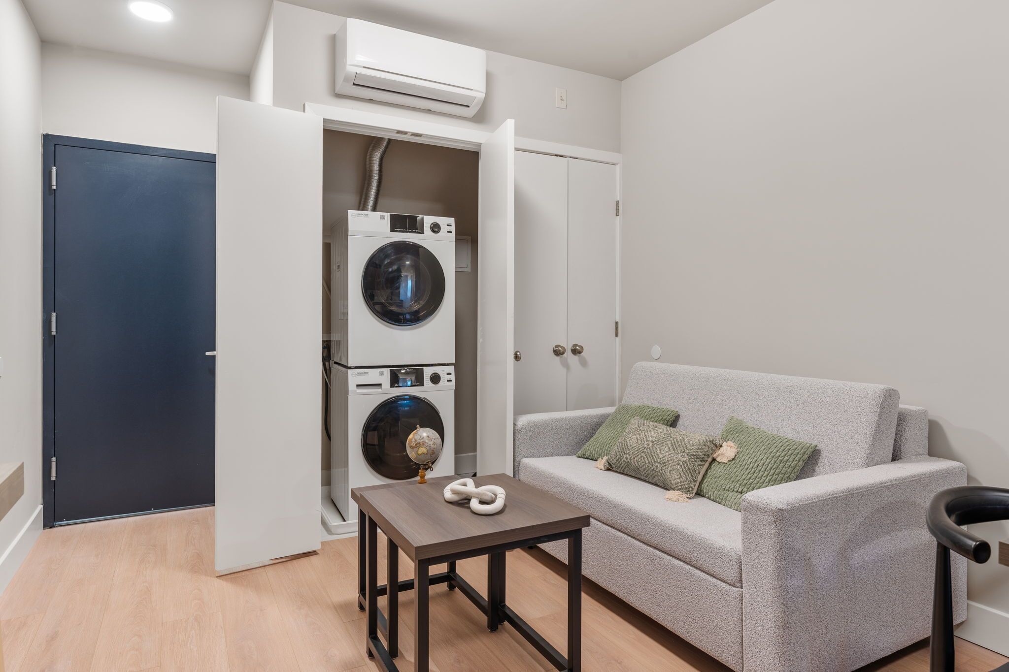 A compact living space with a grey sofa, a small table, a washing machine, and a dark blue door.