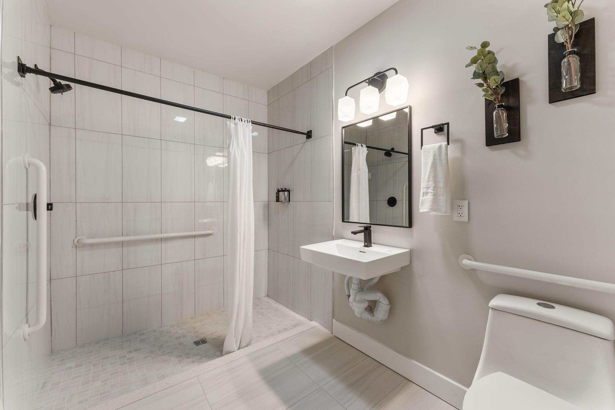 A modern bathroom with a glass shower enclosure, a white sink, a mirror with lights, and two wall-mounted planters.