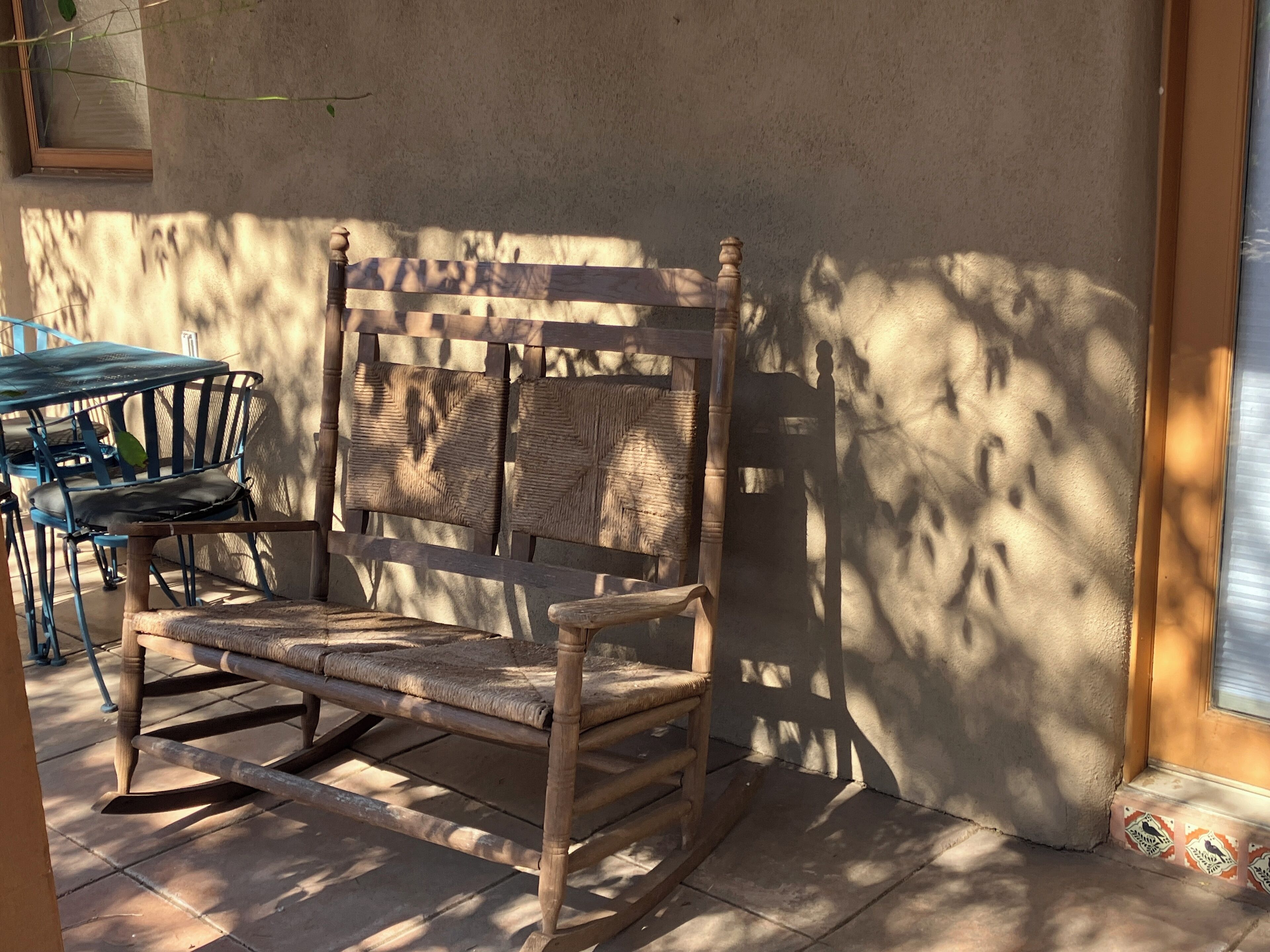 Una mecedora de madera con asiento y respaldo tejido, ubicada frente a una pared con ventana.