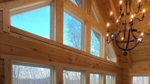 Un vaste espace dédié à la salle à manger, avec une grande table en bois et six chaises, de grandes fenêtres offrant une vue sur une terrasse et les arbres, et un lustre suspendu au plafond.