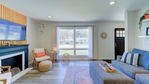 A living room with a fireplace, a television, a sofa, and a large window with a view of the outside. - Walk to Beach, Ping Pong, King Beds, Sleeps 6 (Muskegon)