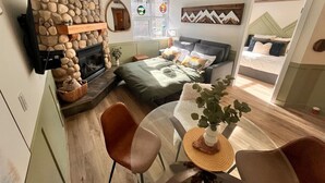 A cozy living room with a stone fireplace, a bed with green bedding, a dining table with chairs, and a large window with a view. - Three Peaks Getaway - sleeps 4 (Canmore)