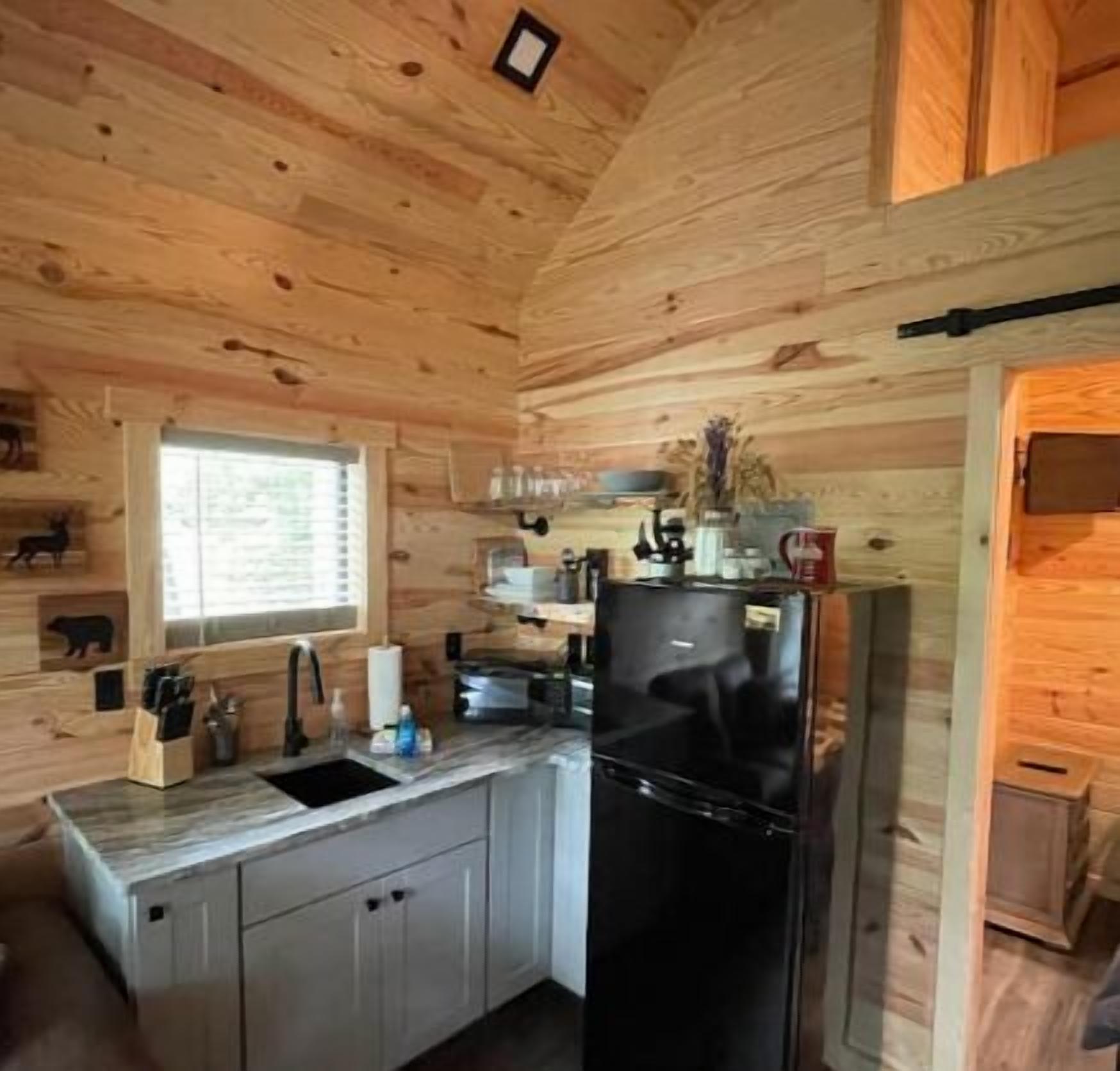 A compact kitchen with wooden walls, a black refrigerator, and a window with blinds.