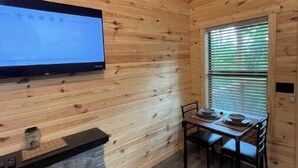 A wooden cabin interior with a TV mounted on the wall, a fireplace, a dining table with chairs, and a window with blinds. - Bear Top Junior Small Cabin w EV Charger

 (Cosby)