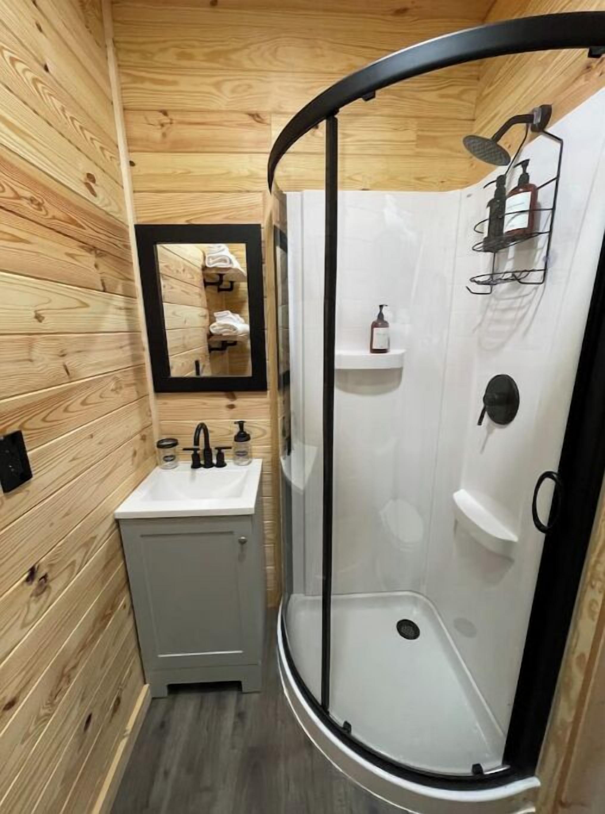 A compact bathroom with a wooden wall, a sink with a mirror, and a glass-enclosed shower area.