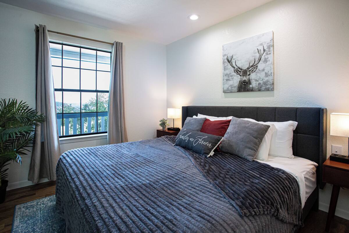 Une chambre à coucher avec un grand lit, une fenêtre avec des rideaux, un tableau représentant un cerf et une table de chevet avec une lampe.