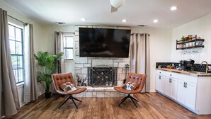 A modern living room with a fireplace, two leather armchairs, a flat-screen TV, and a kitchen area with white cabinets and a countertop. - Hot Tub Retreat by Wine Country (Fredericksburg)