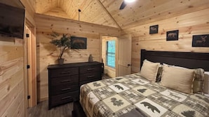 Une chambre dans un chalet en bois, avec un lit, une commode, un ventilateur de plafond et un téléviseur fixé au mur.
