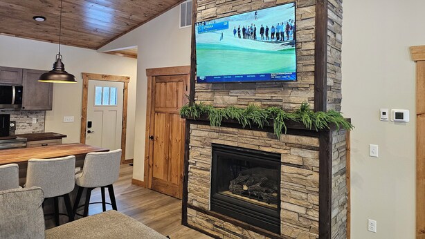 A modern kitchen with a stone fireplace, a dining area with bar stools, and a television mounted on the wall displaying a golf course. - NEW 3-bedroom cabin in beautiful Emily, MN.  Located right on the trails! (Emily)