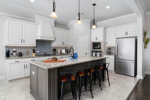 Une cuisine moderne avec des meubles blancs, un grand îlot central doté de tabourets, des appareils en acier inoxydable et un éclairage suspendu.