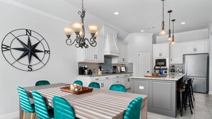 A modern kitchen with white cabinets, a central island, and a dining area with teal chairs. - The Noble House Near Magnolia with Pool (Hewitt)