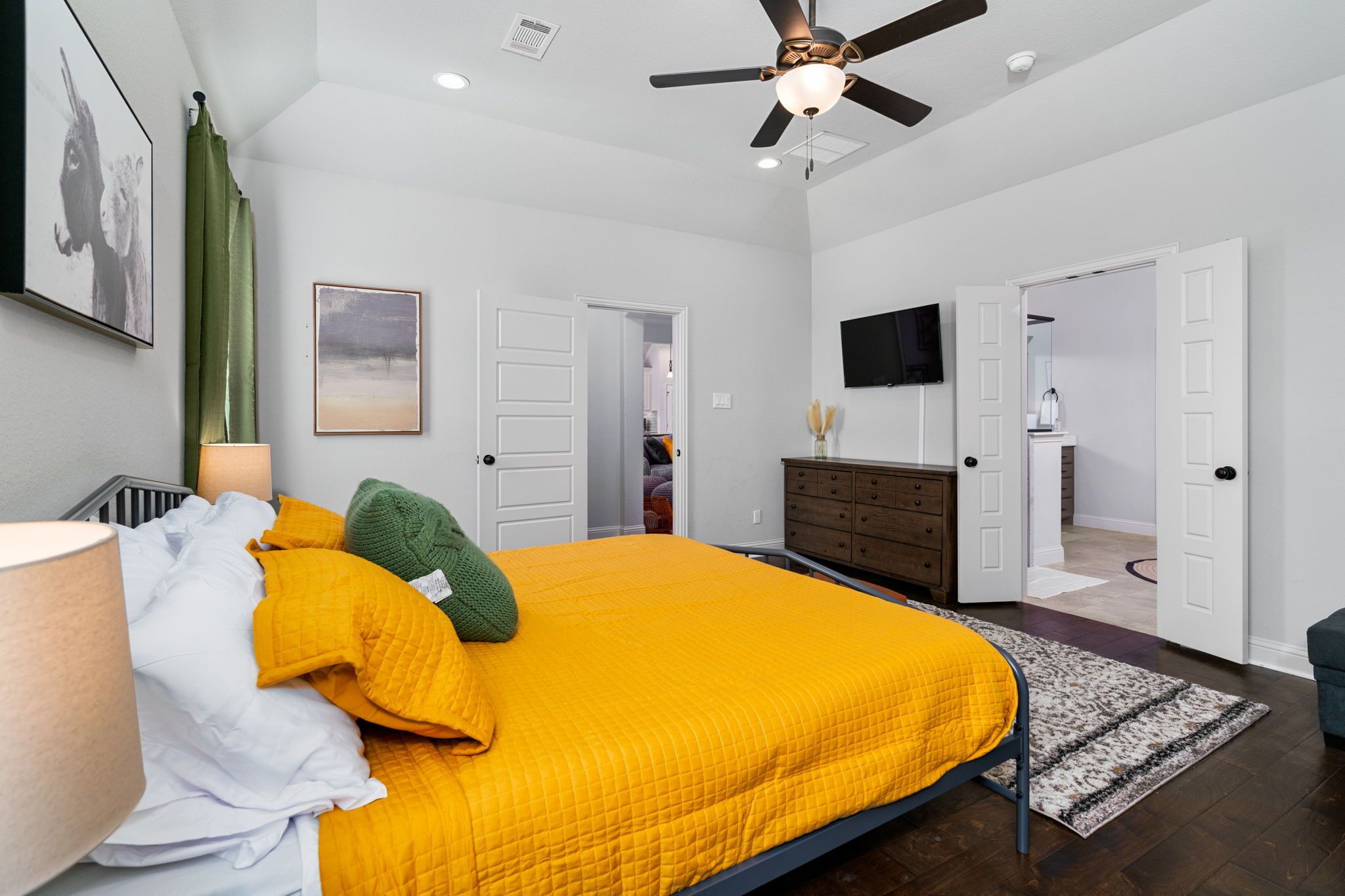 A bedroom with a large bed, a ceiling fan, a dresser, and a television on the wall.