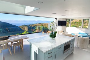 A modern kitchen with a large island, wooden chairs, and a view of a hilly landscape.