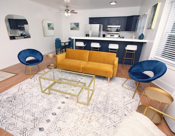 Ein modernes Wohnzimmer mit einem gelben Sofa, blauen Sesseln, einem Couchtisch und einer Küche im Hintergrund.