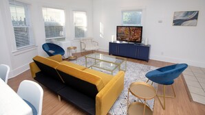 A modern living room with a yellow sofa, blue chairs, a glass coffee table, and a television on a blue cabinet.