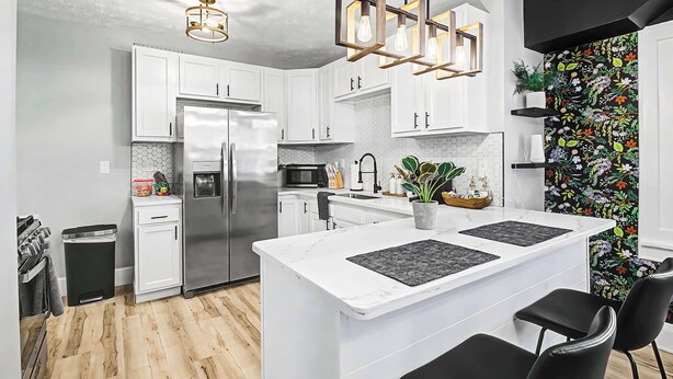 A modern kitchen with white cabinetry, a stainless steel refrigerator, a central island with black mats, and a black and white floral wallpaper accent wall.