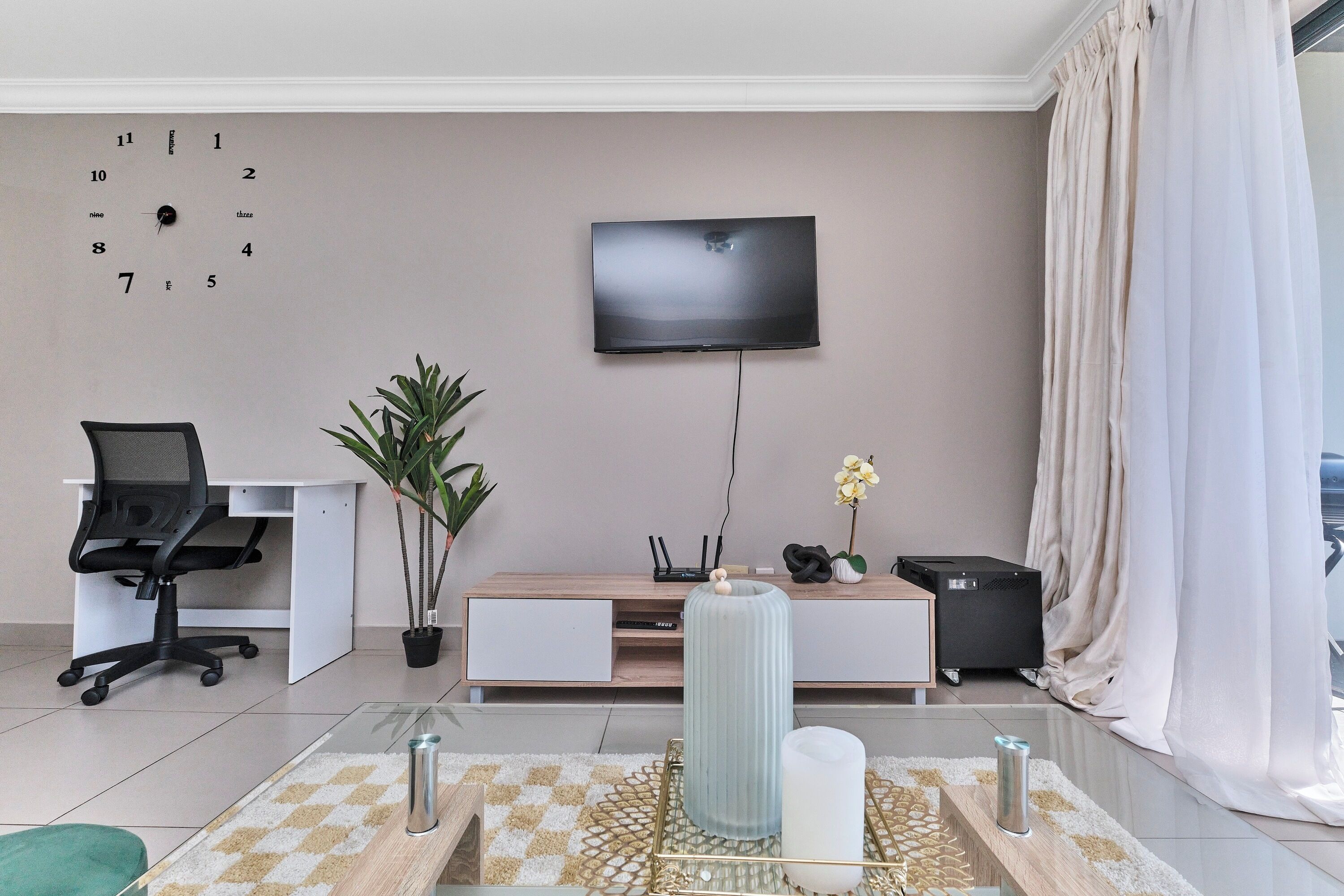 Un salon moderne avec un téléviseur à écran plat fixé au mur, une table basse en bois agrémentée d’objets décoratifs, un fauteuil de bureau noir, une plante en pot et un rideau blanc.
