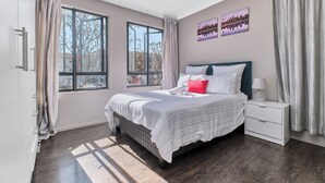 A bedroom with a large bed, a nightstand, and two windows. - Luxury & Secure apartment - Fourways (Fourways)