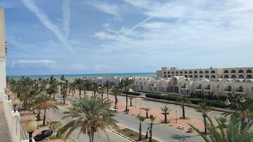 A coastal hotel with a view of the sea, palm trees, and a street with parked cars.