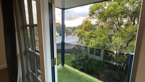 Uitzicht vanaf een balkon met een glazen deur, op een gazon met bomen en een gebouw in de verte.