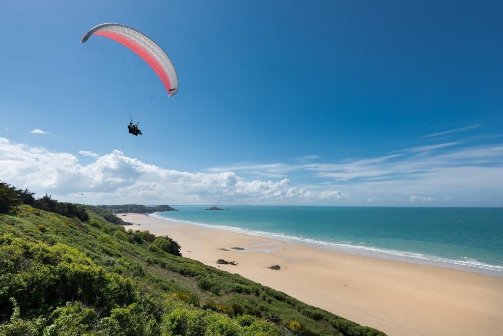 Une plage où l’on voit une personne faire du parapente, un ciel dégagé et des reliefs lointains.