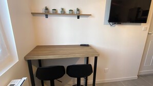 Een compacte kitchenette met een houten tafel en twee zwarte krukken, een plank met potplanten, en een wandgemonteerde flatscreen-tv.