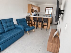 A modern living space with a blue sofa, a dining area with bar stools, a kitchen with wooden cabinets, and a flat-screen TV mounted on the wall.