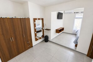A bedroom with a wooden wardrobe, a mirror with lights, a small shelf, a trash bin, and a bed.