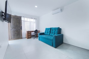 Ein Zimmer mit einem blauen Sofa, einem Holztisch, einem Stuhl, einem an der Wand befestigten Fernseher und einem Fenster mit Vorhängen.