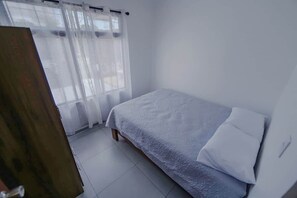 A small, single-bed room with a wooden door, a white bed with a textured blanket, and sheer curtains on the window. - Casa Monarch La Fortuna • Pool • Volcano Views (La Fortuna)