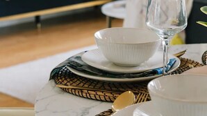 Une table à manger dressée avec des bols blancs et un verre à vin, un set de table en fibres tressées et une cuillère dorée.