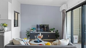 Un salon moderne avec un canapé gris, une petite table ronde et un téléviseur posé sur un meuble noir. On y trouve également des plantes en pot et une grande fenêtre avec des rideaux.