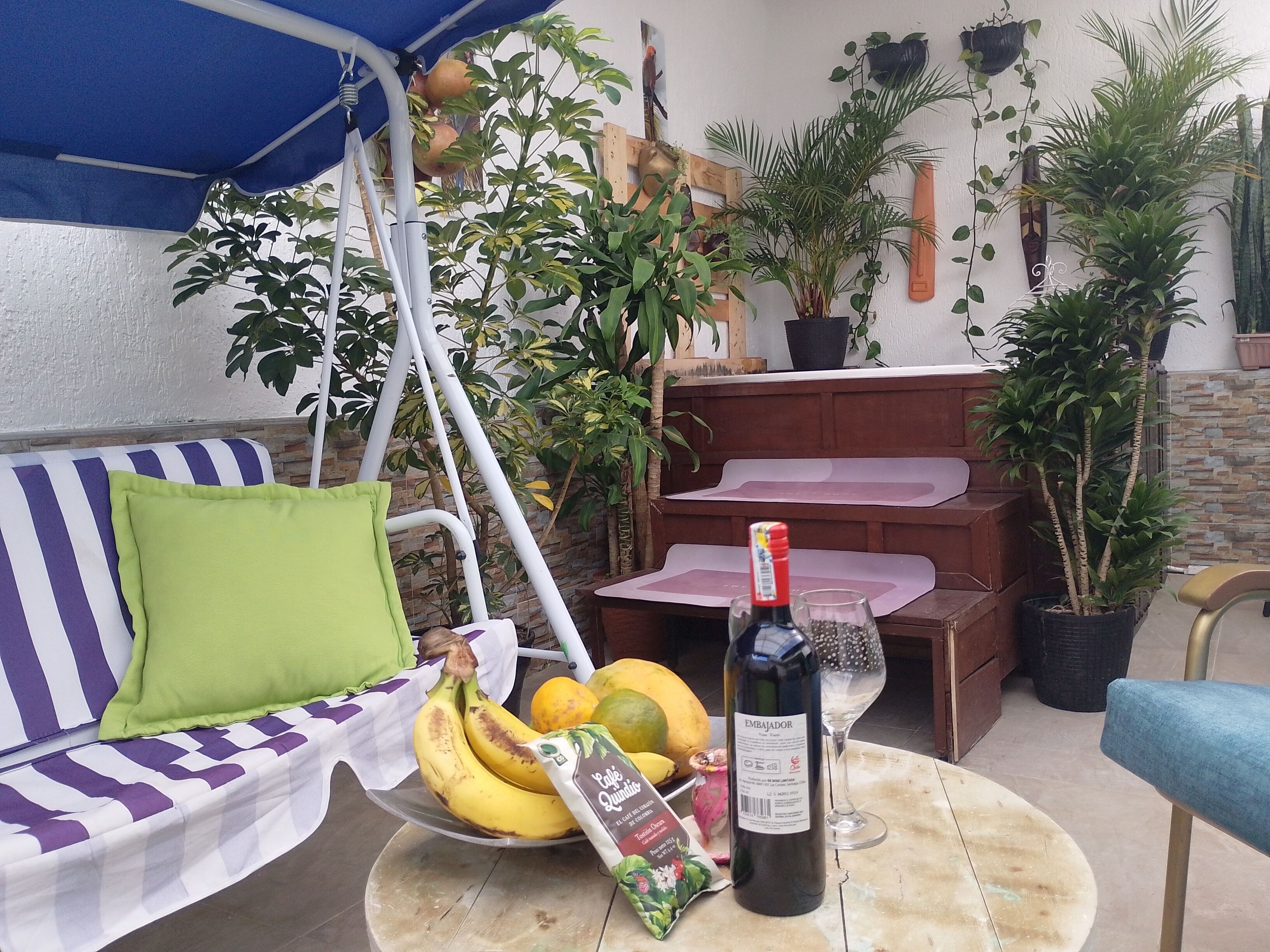 En uteplats med en randig soffa, en hängstol, ett bord med en vinflaska, ett glas och en skål med frukt.