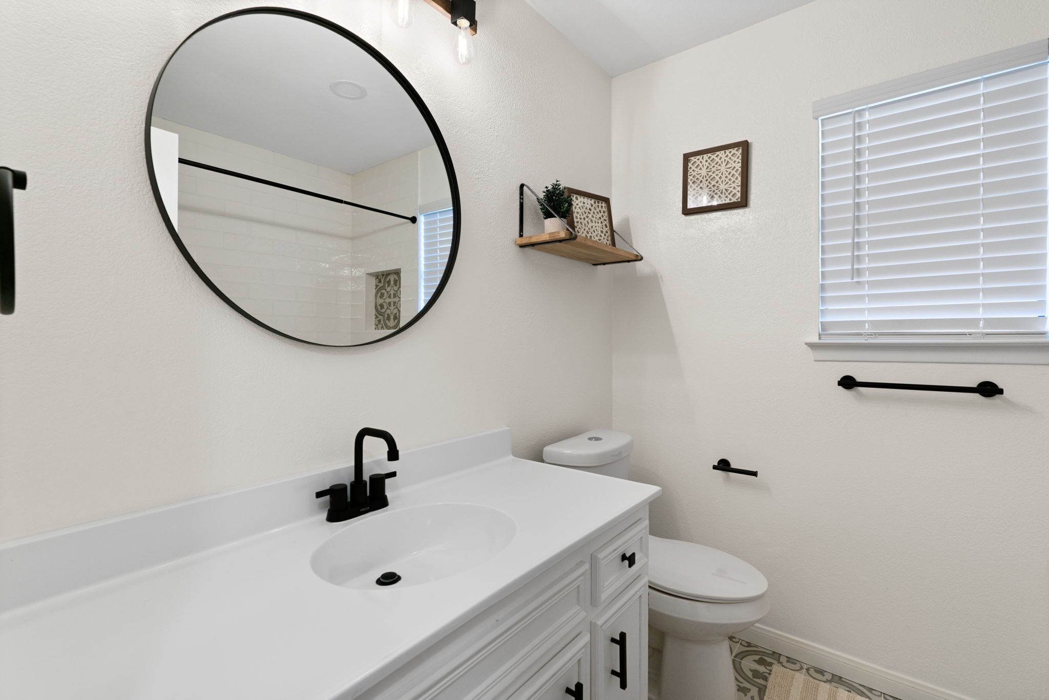 Un baño con un lavabo blanco, un grifo negro y un espejo redondo. Hay un inodoro con tapa blanca y un pequeño estante encima.
