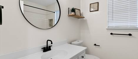 Un baño con un lavabo blanco, un grifo negro y un espejo redondo. Hay un inodoro con tapa blanca y un pequeño estante encima.